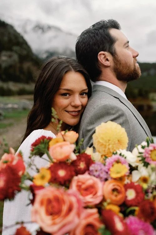 A bride leans onto the shoulder o her new husband while holding her colorful bouquet by a mountain lake