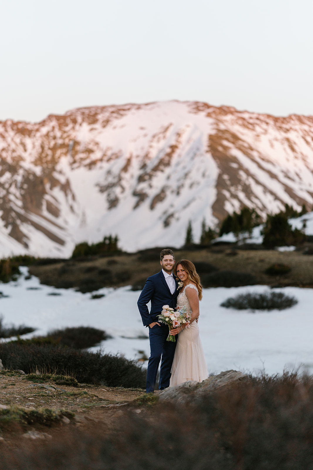 Newlyweds stand together in a mountain trail in front of snow-covered mountains