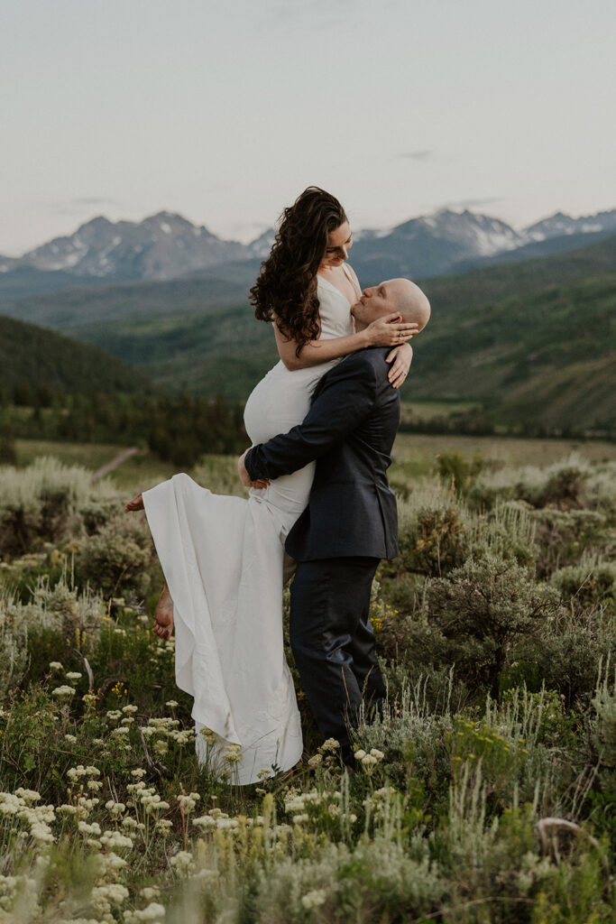 Groom lifts the bride joyfully in the mountain meadow after their private elopement toast.