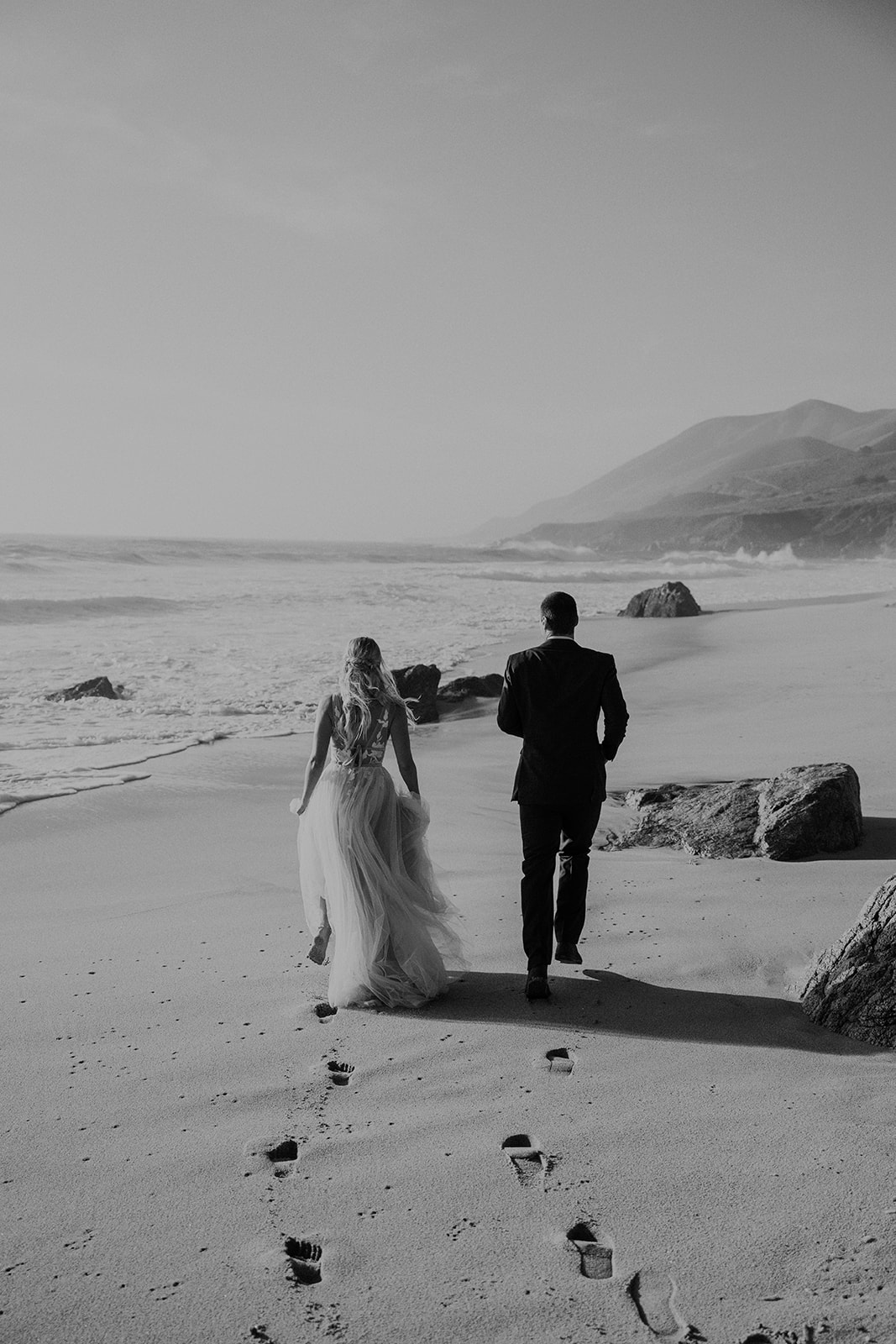Black and white photo of a bride and groom running along the beach in Big Sur