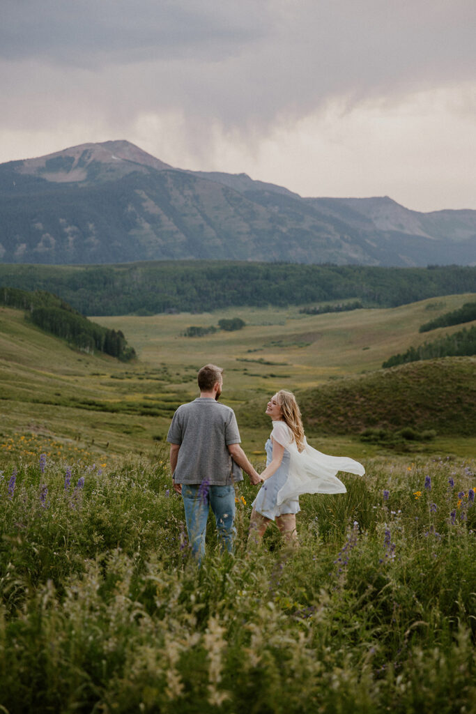 Couple holding hands and walking through wildflower fields in Crested Butte with mountain views in the background.