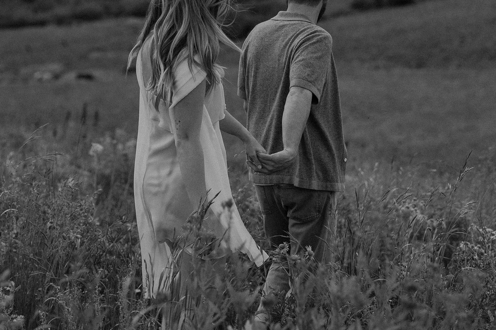 Black and white photo of couple walking away hand-in-hand through tall grass during Crested Butte engagement session.