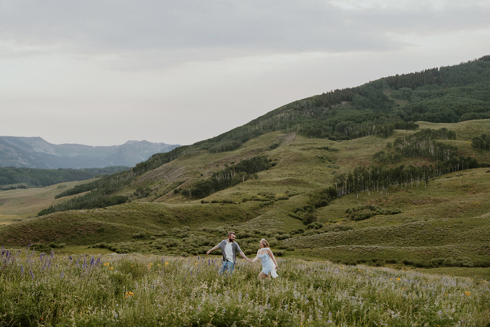 Wide shot of couple running hand-in-hand through a valley of wildflowers with the Crested Butte mountains in the distance.