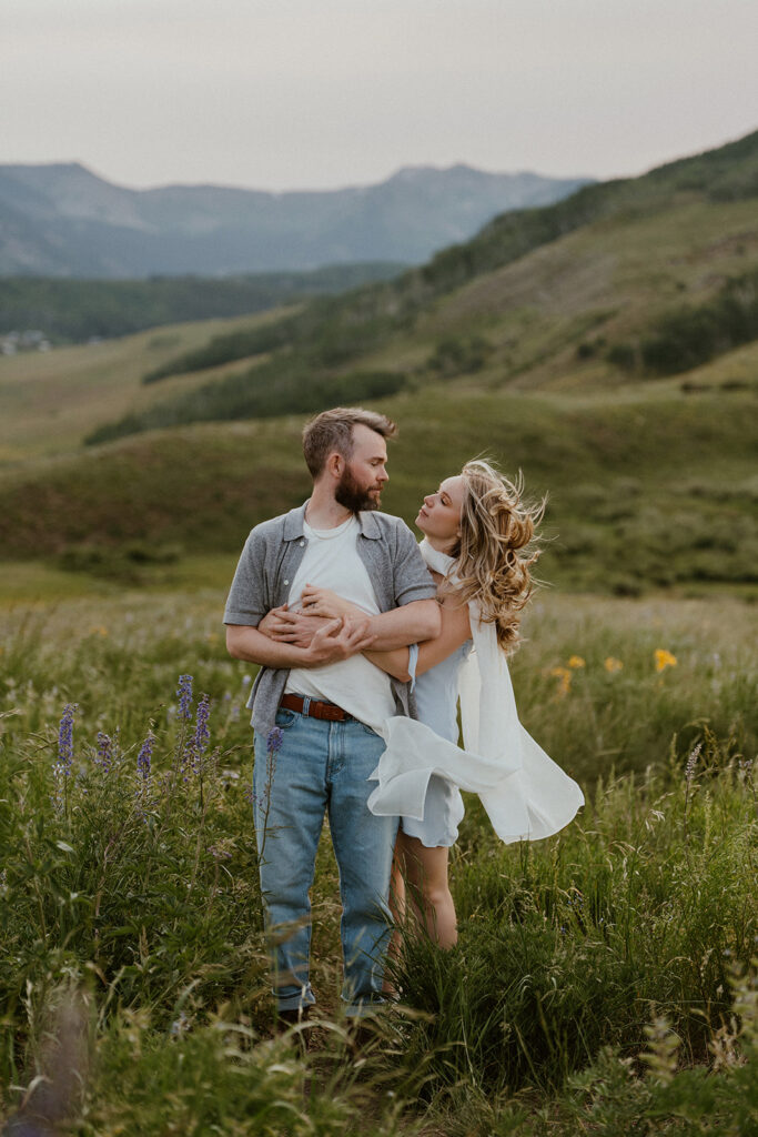Couple embracing in a meadow, surrounded by purple and yellow wildflowers with green mountains in the background.