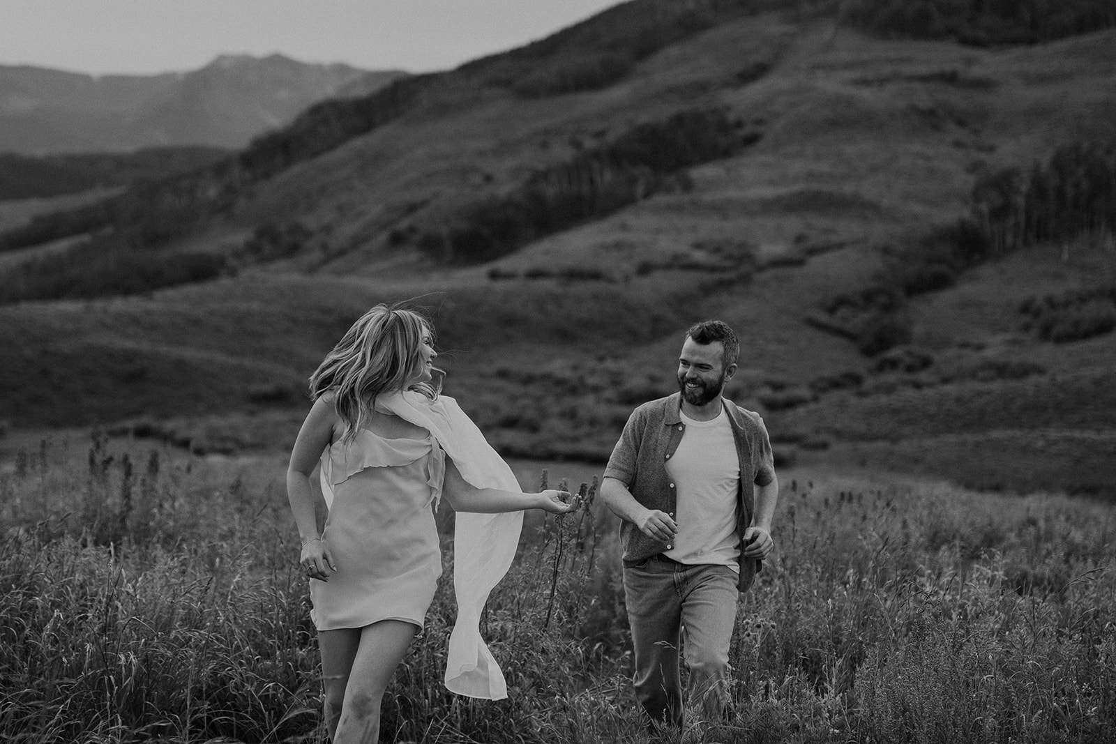 Black and white wide shot of couple running hand-in-hand through a valley of wildflowers with the Crested Butte mountains in the distance.