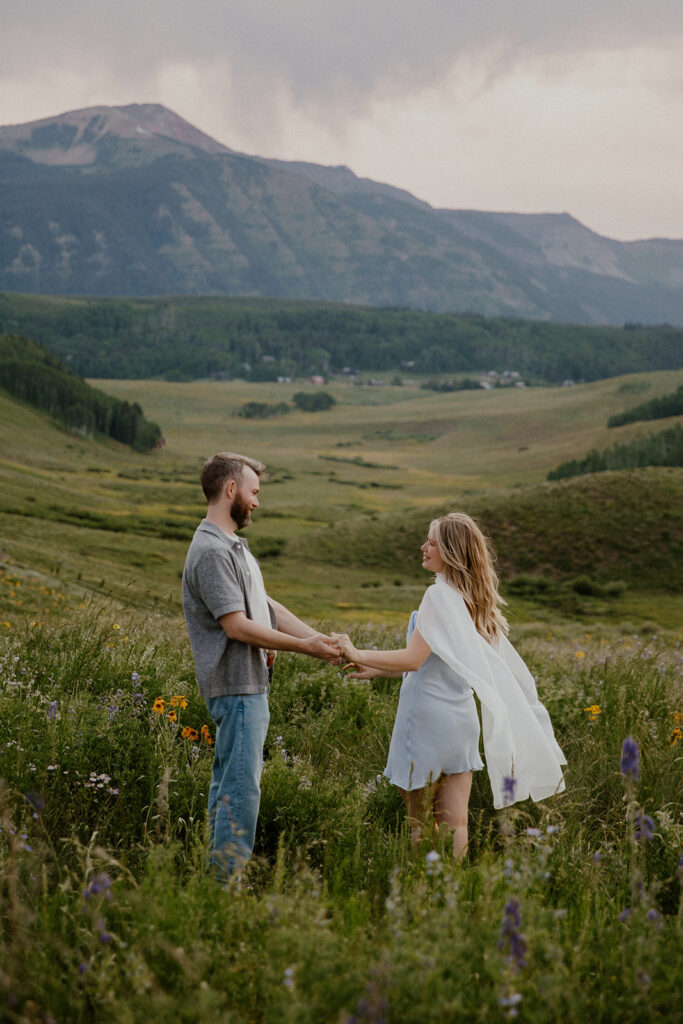 Couple standing and holding hands in a wide meadow with mountains behind them during their Crested Butte engagement session.