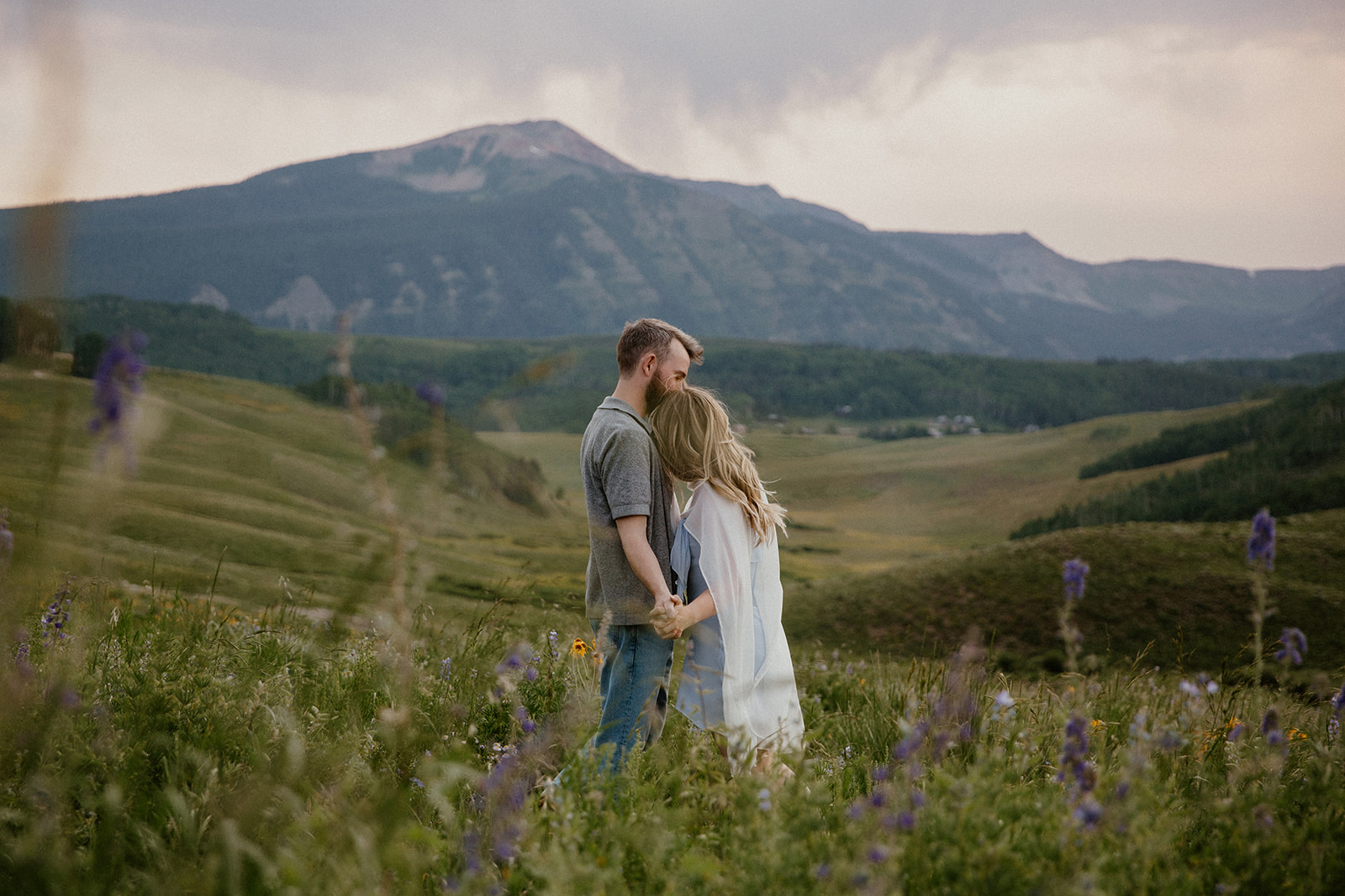 Couple standing face to face, holding hands in a wide meadow with mountains behind them during their Crested Butte engagement session.