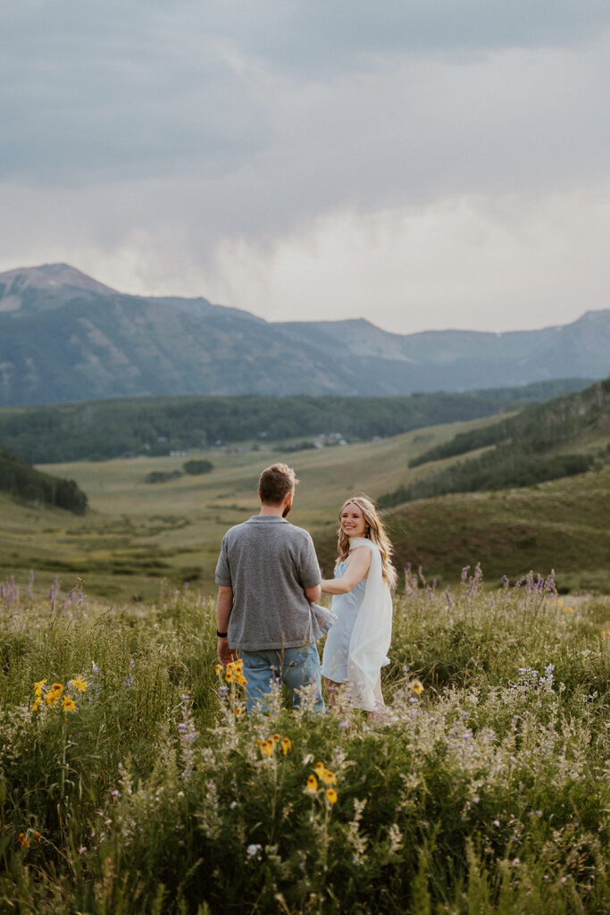 Woman looking back and smiling as she walks hand-in-hand with her fiancé in a colorful field of wildflowers.