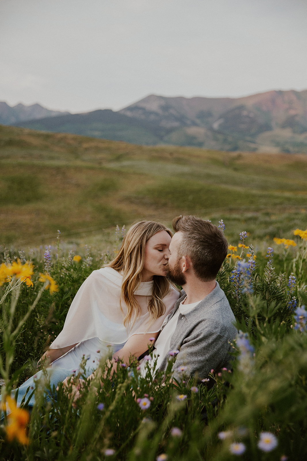 Intimate moment as the couple shares a kiss in the field, surrounded by tall grass and wildflowers with mountain views.