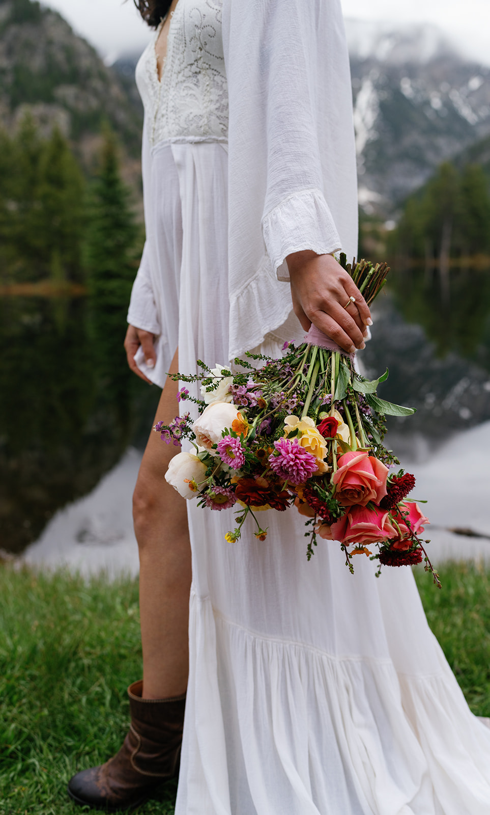 A bride in boots and a white dress holds her colorful bouquet while walking by a mountain lake on green grass