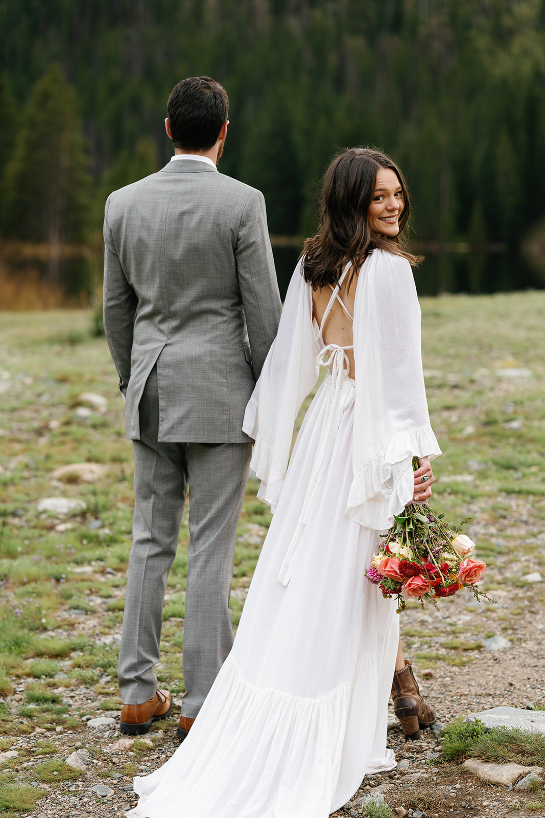 A bride looks over her shoulder while holding her husband's hand in a mountain trail