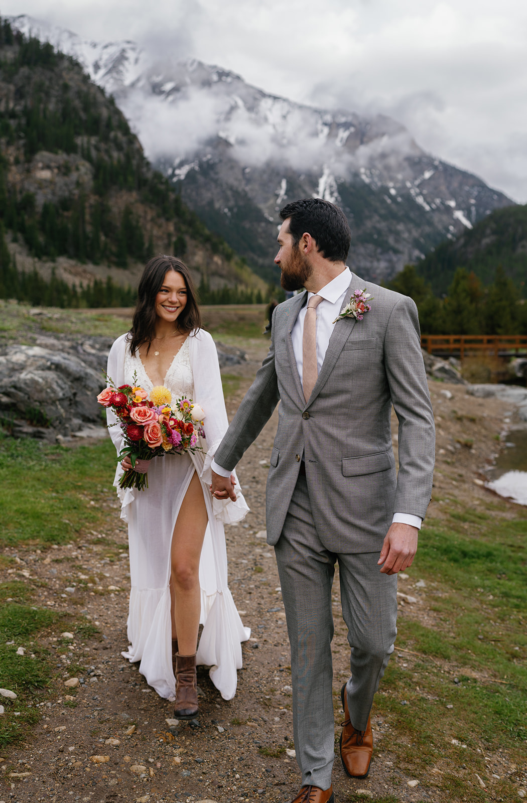 A groom leads his new bride down a mountain trail by the hand while she holds her large bouquet devils thumb ranch wedding