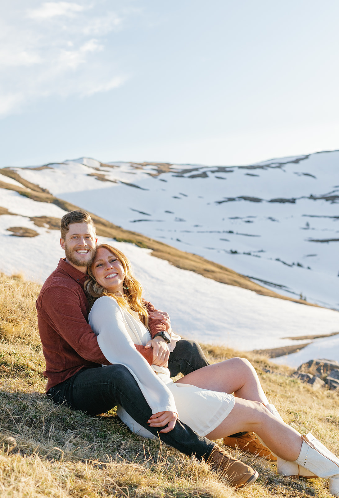 A woman in a white dress sits in the lap of her partner wearing a red shirt and dark jeans on a snowy mountainside