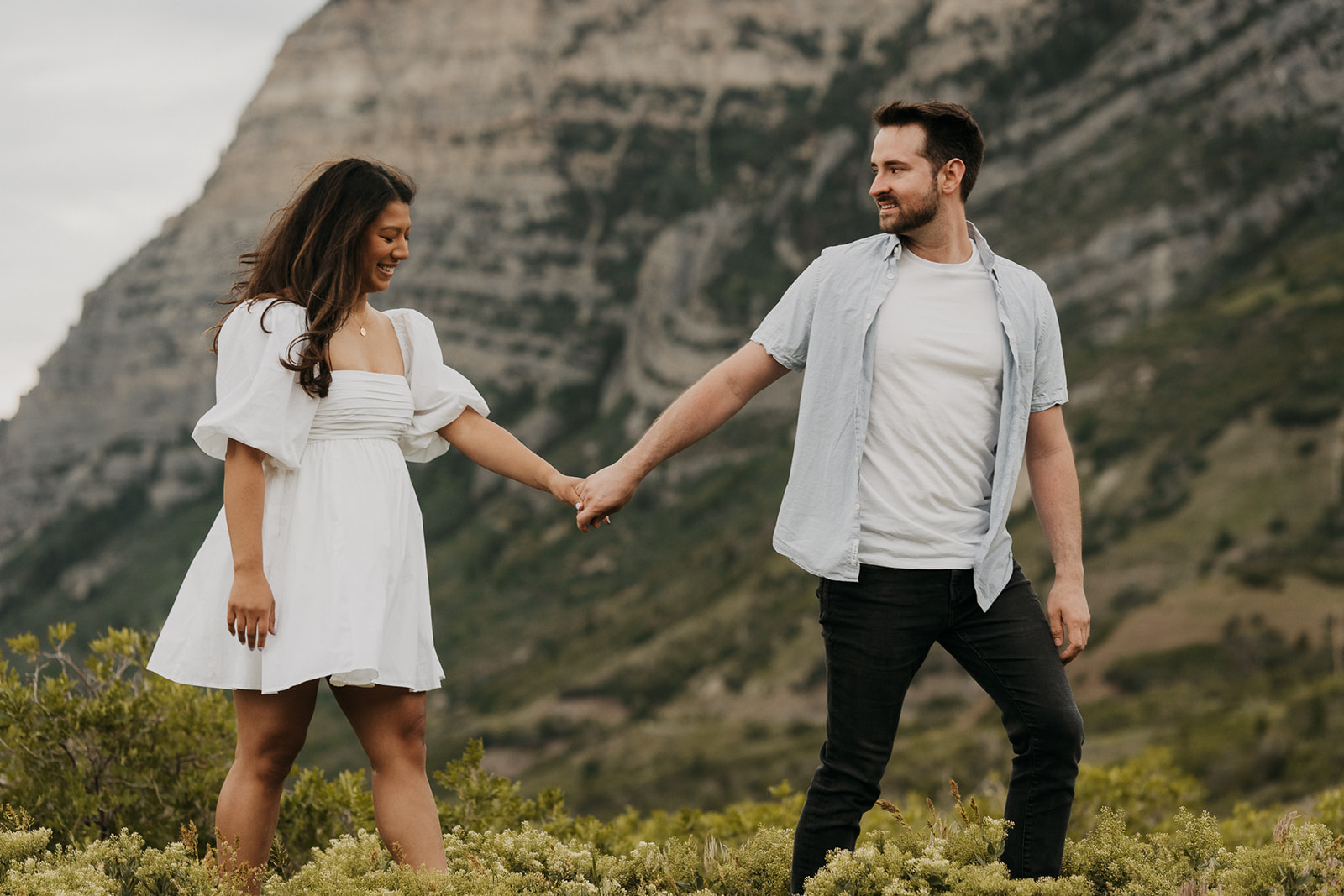 A man leads his fiancee through a mountain trail while holding hands in a white dress