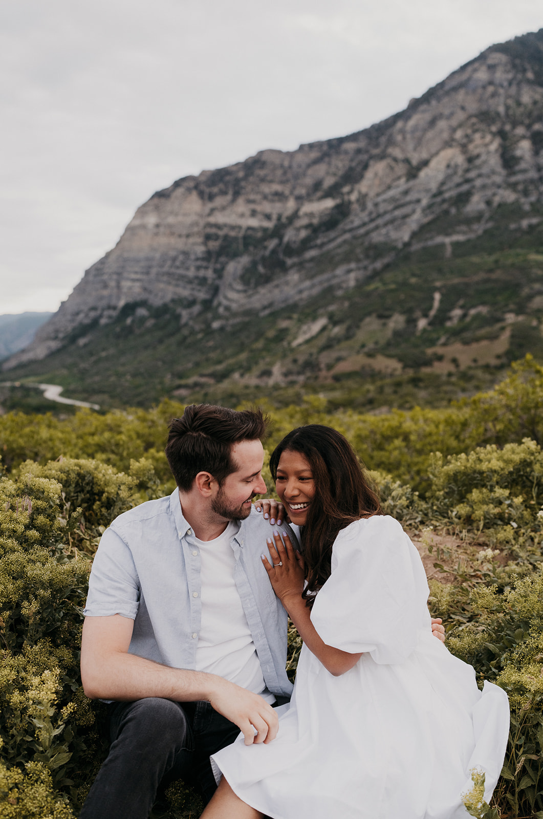 A couple sit and laugh together in a mountain trail in a white dress and blue shirt Donovan Pavilion wedding