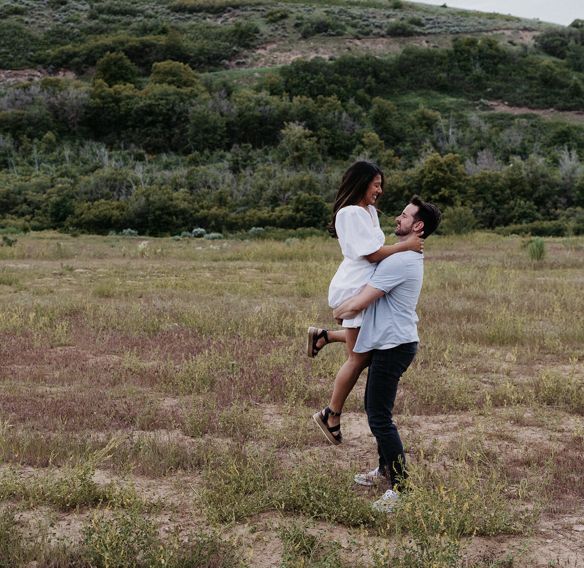 A man lifts his bride while standing in a field by a large hill