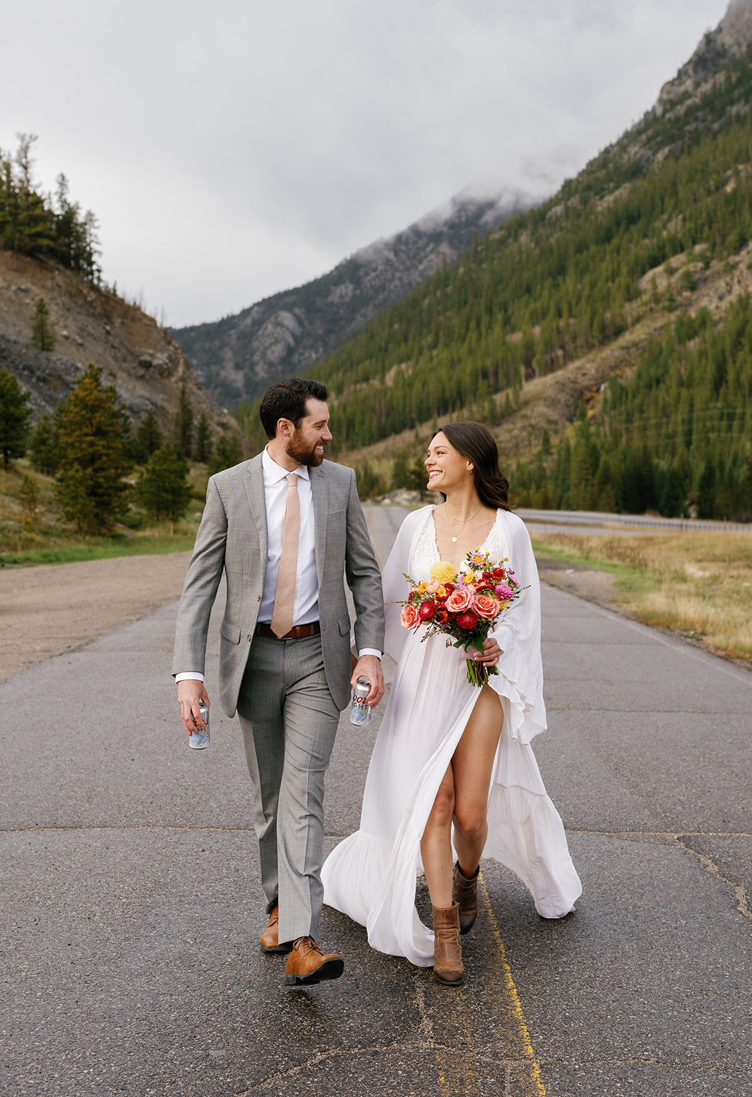 Newlyweds walk down a road in the mountains holding beer and flowers Keystone Resort Wedding
