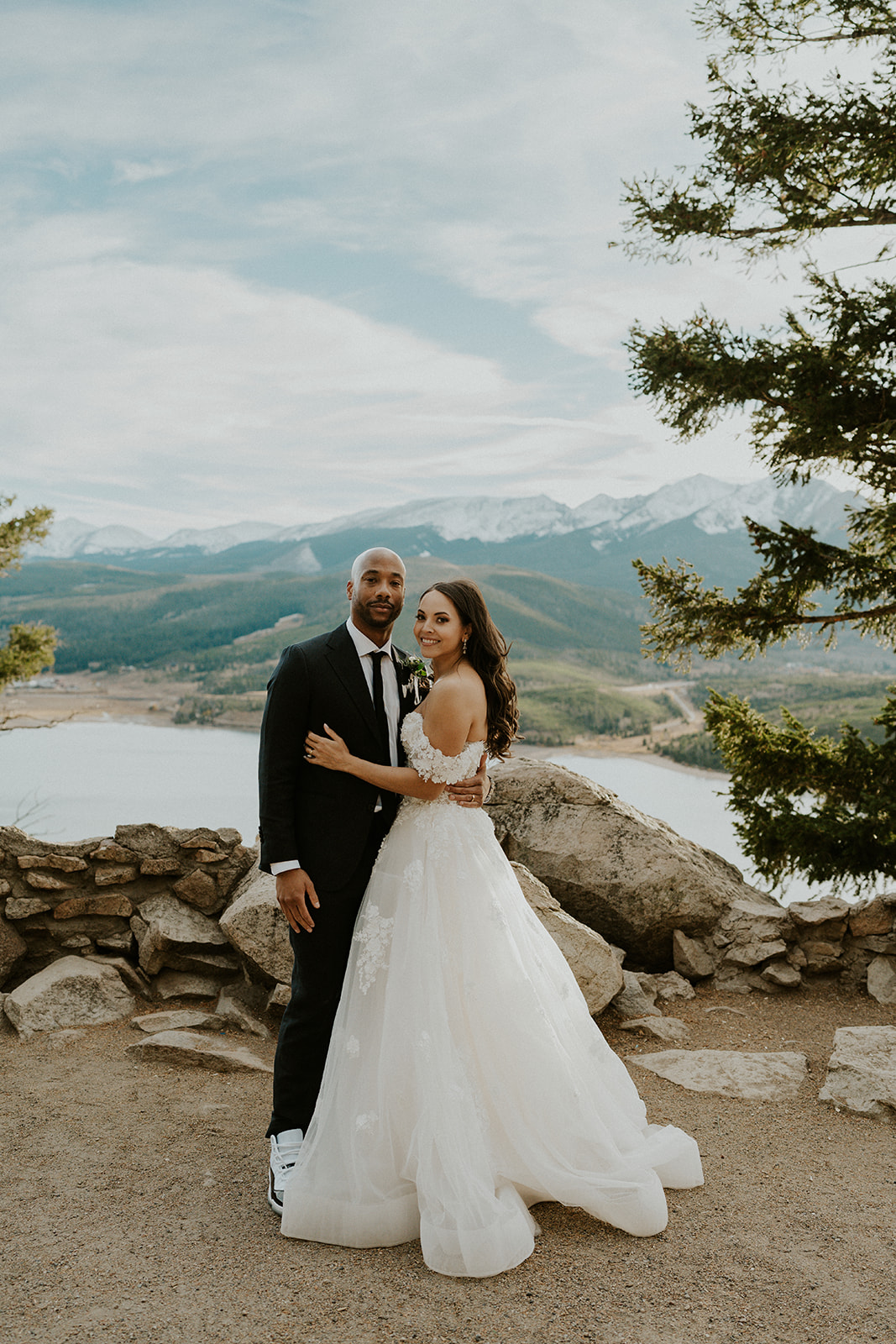 Newlyweds stand together in a mountain trail overlooking a lake at sunset