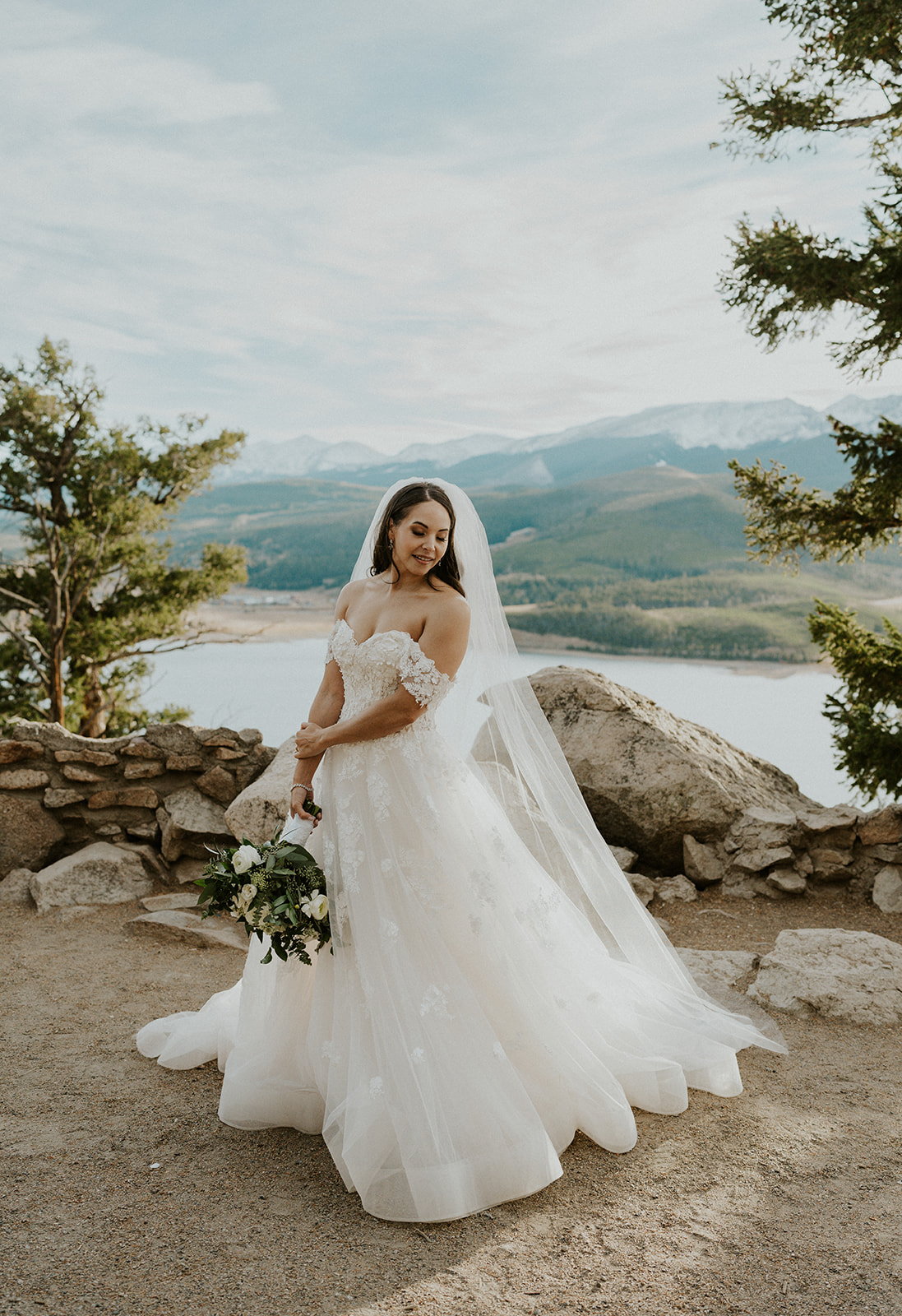 A bride looks back over her shoulder while standing on the edge of the mountain overlooking a lake at her Cottonwood Pass elopement