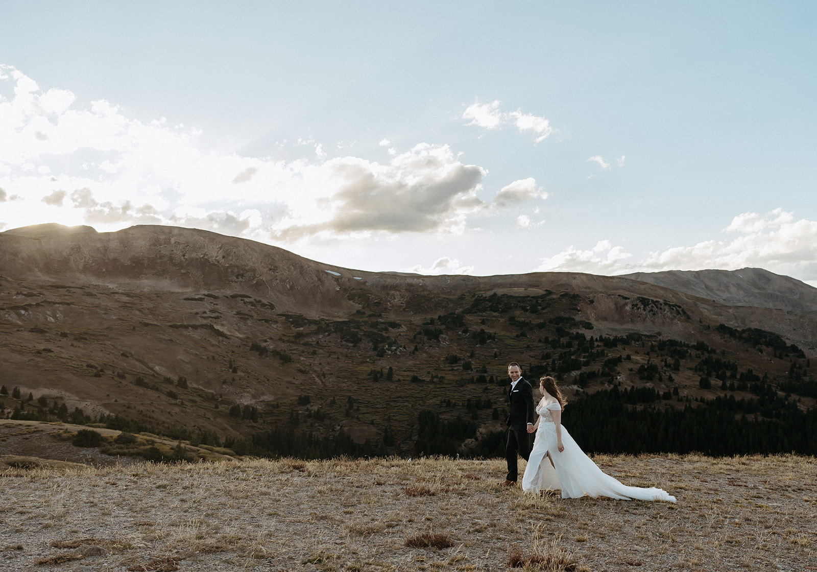 Newlyweds hold hands while walking through a remote mountain trail at sunset