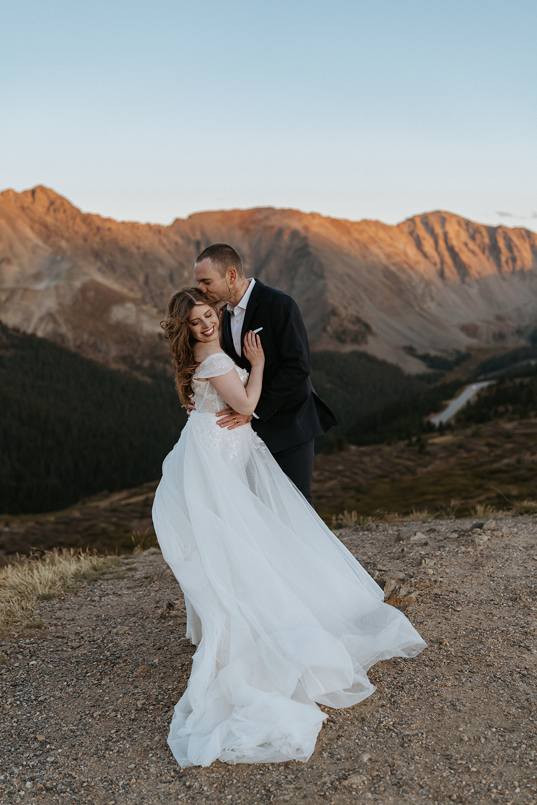 A groom in a black suit kisses the temple of his bride in a lace dress at sunset in the remote mountains