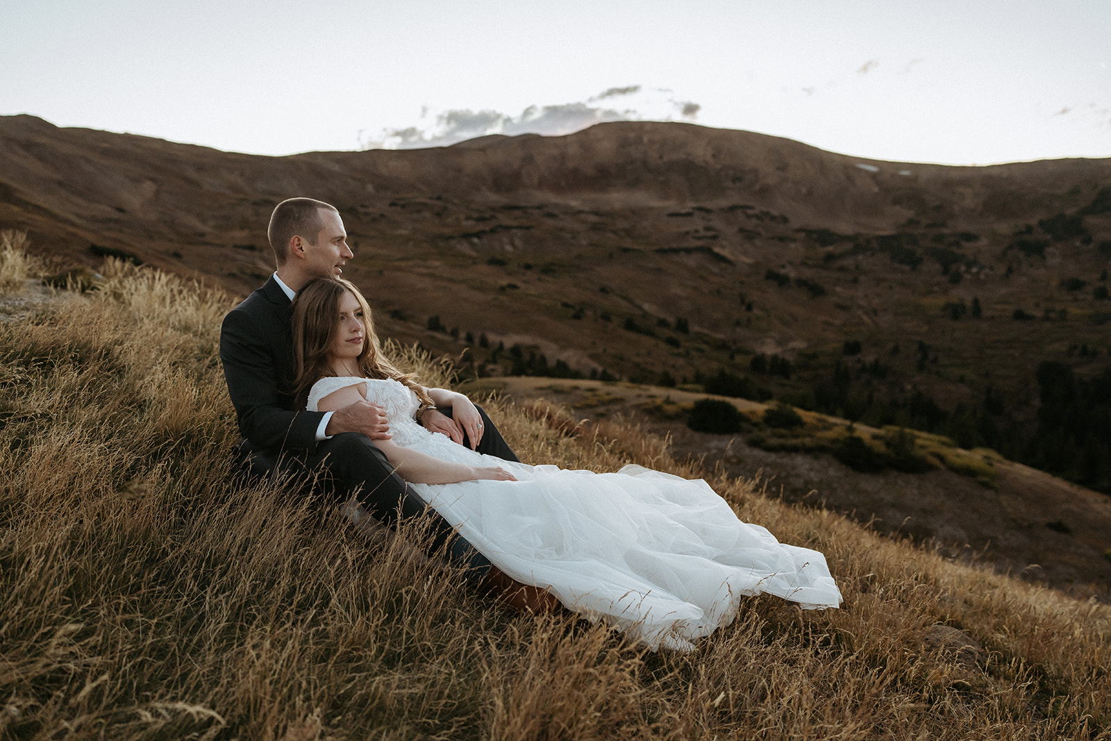 A bride lays in the lap of her husband on a steep hillside at sunset