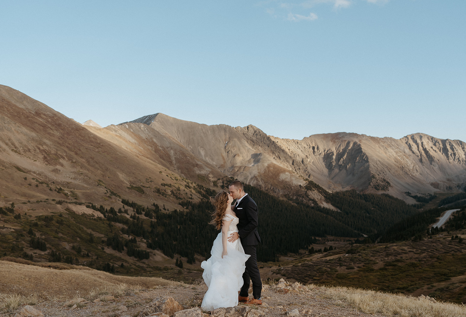 Newlyweds kiss on the edge of a mountain at sunset in a lace dress and black suit
