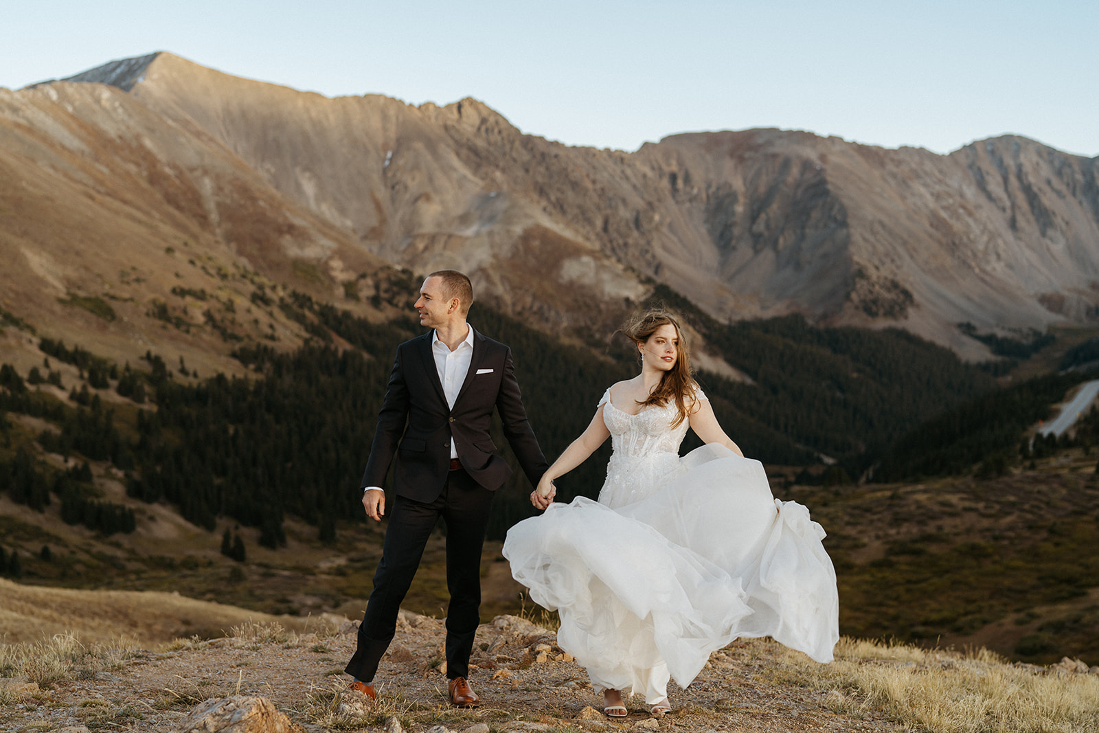 A bride plays in her lace dress with the wind while holding hands with her groom and standing in the remote mountains