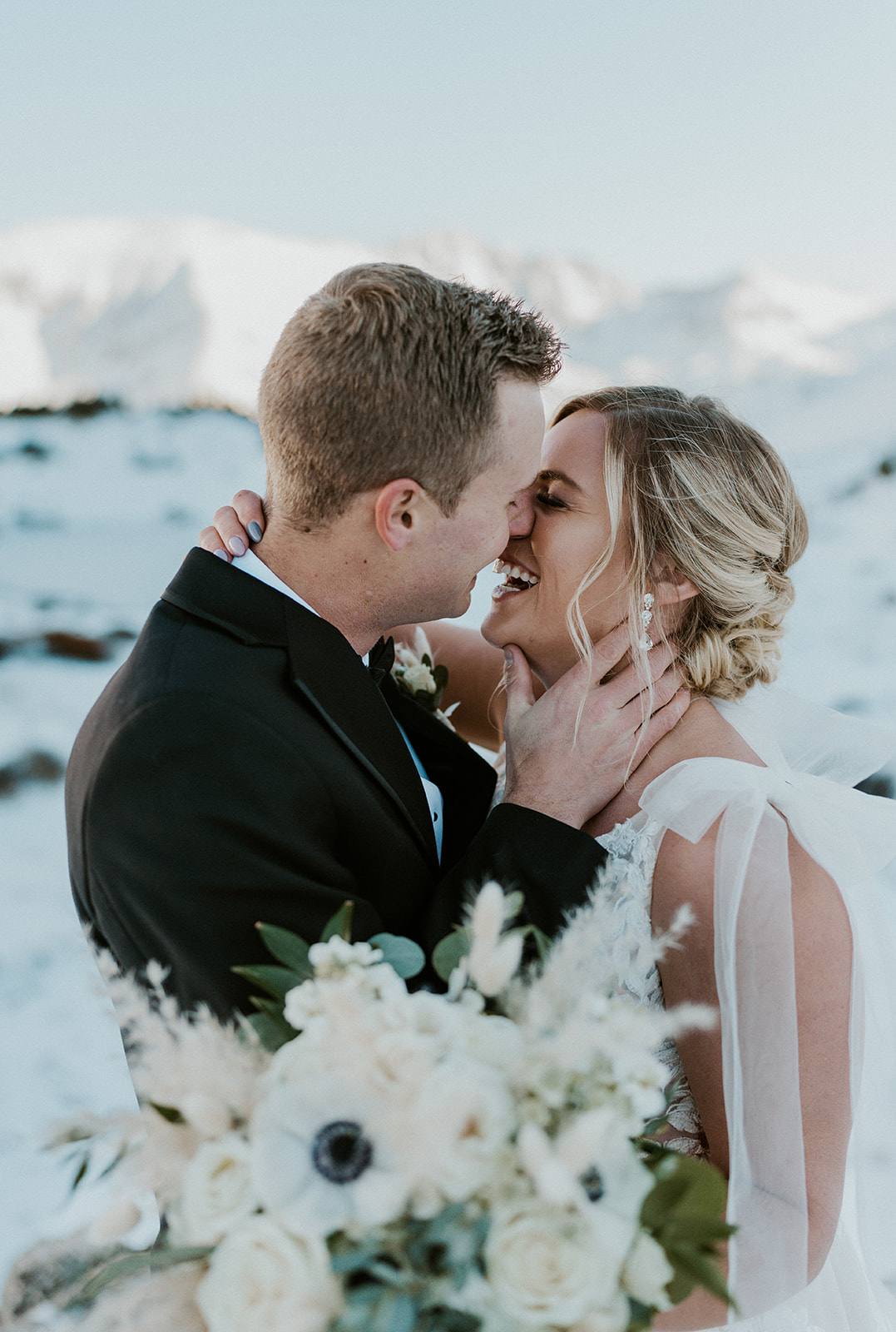 Newlyweds laugh their way to a kiss while standing in a snowy mountain trail