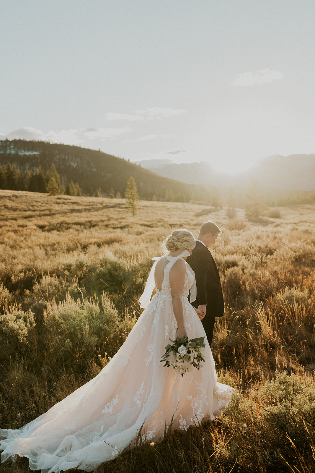 Newlyweds walk hand in hand down a trail through a field of tall grasses at sunset for their Loveland Pass Elopement
