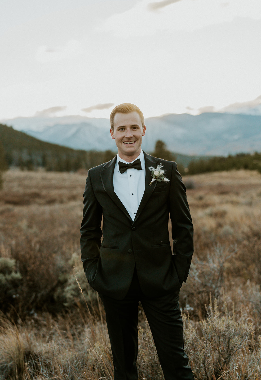 A groom in a black suit stands smiling with hands in his pockets at sunset for his Loveland Pass Elopement