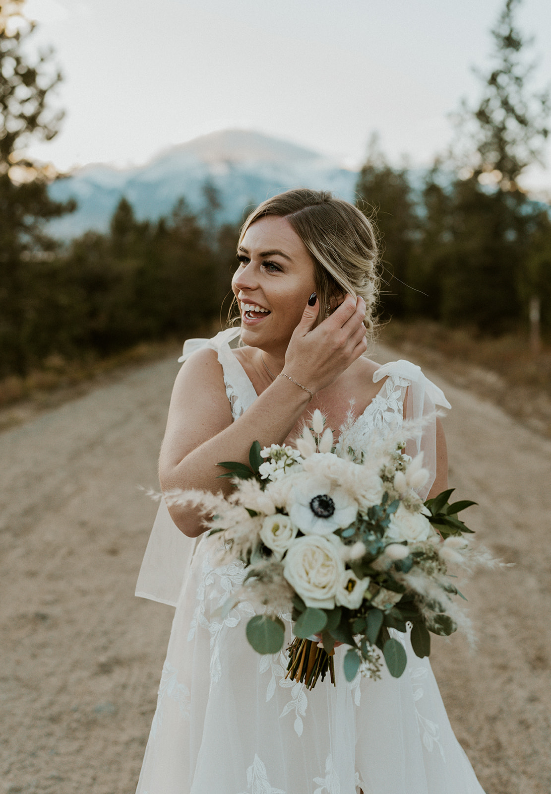 A bride in a cream floral dress stands in a dirt road fixing her hair and holding her white bouquet at her Loveland Pass Elopement