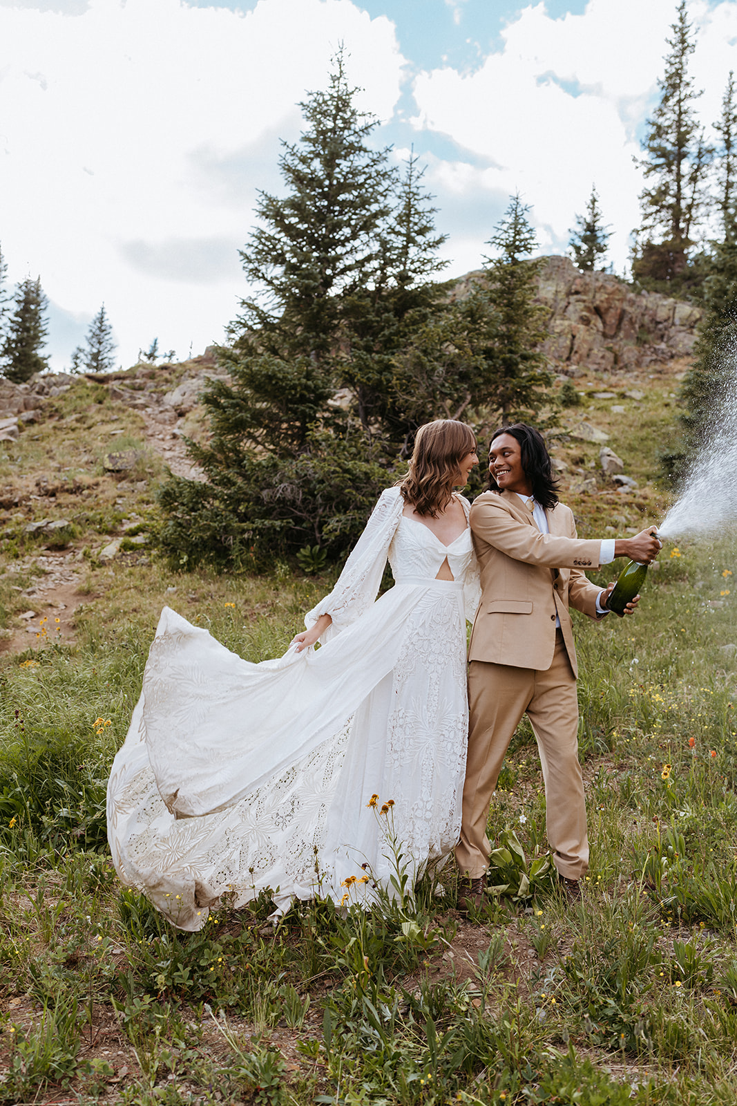 Newlyweds pop a bottle of champagne on a mountainside while smiling at each other