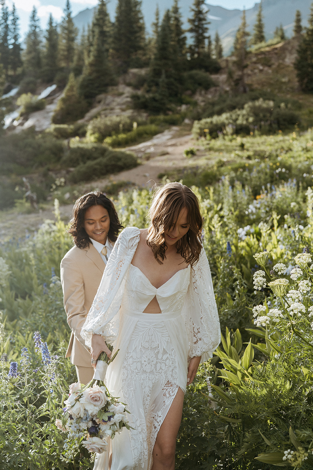 Newlyweds walk up a mountain trail surrounded by wildflowers at sunset