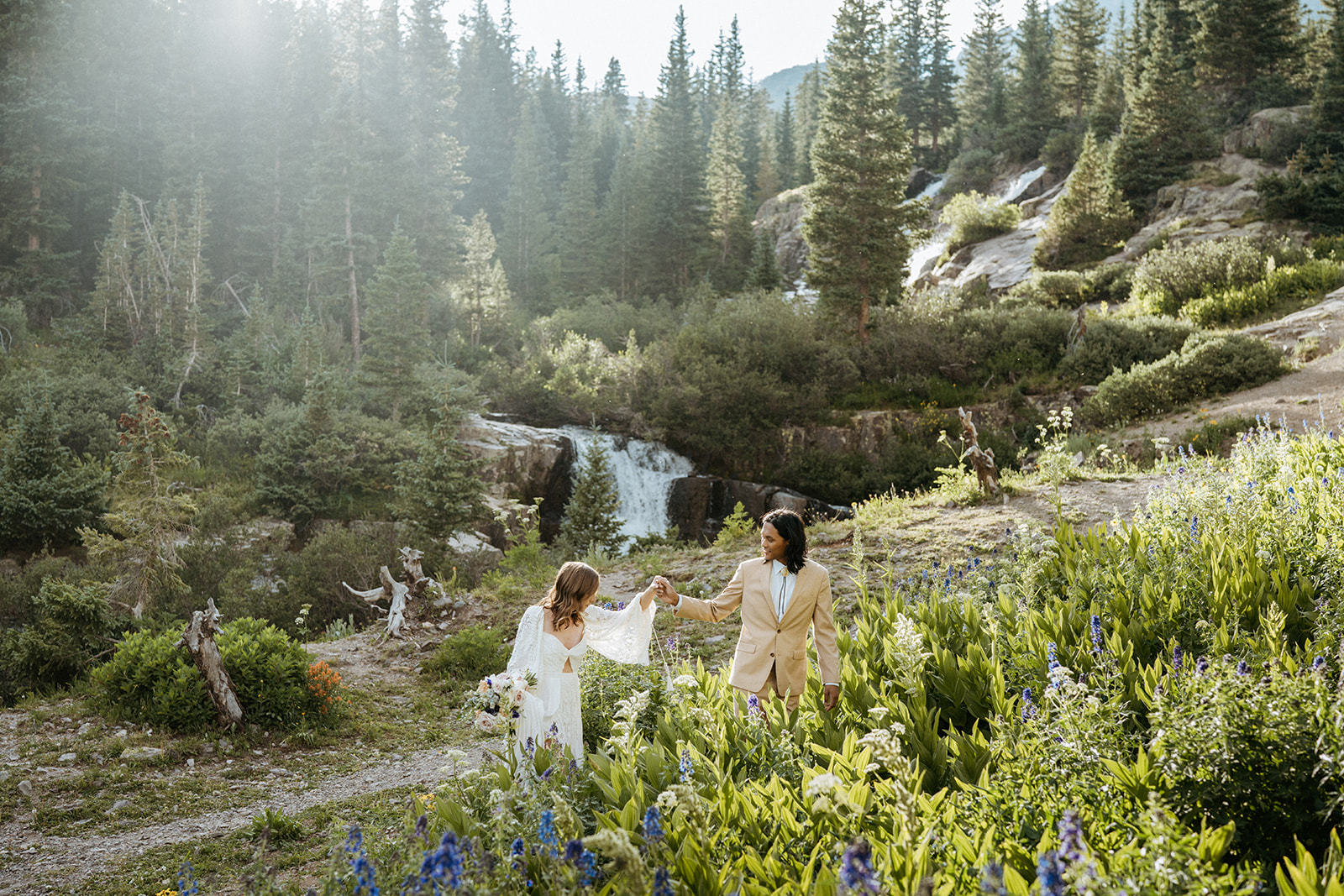 Newlyweds walk up a mountain trail alongside waterfalls at their Ouray Elopement holding hands