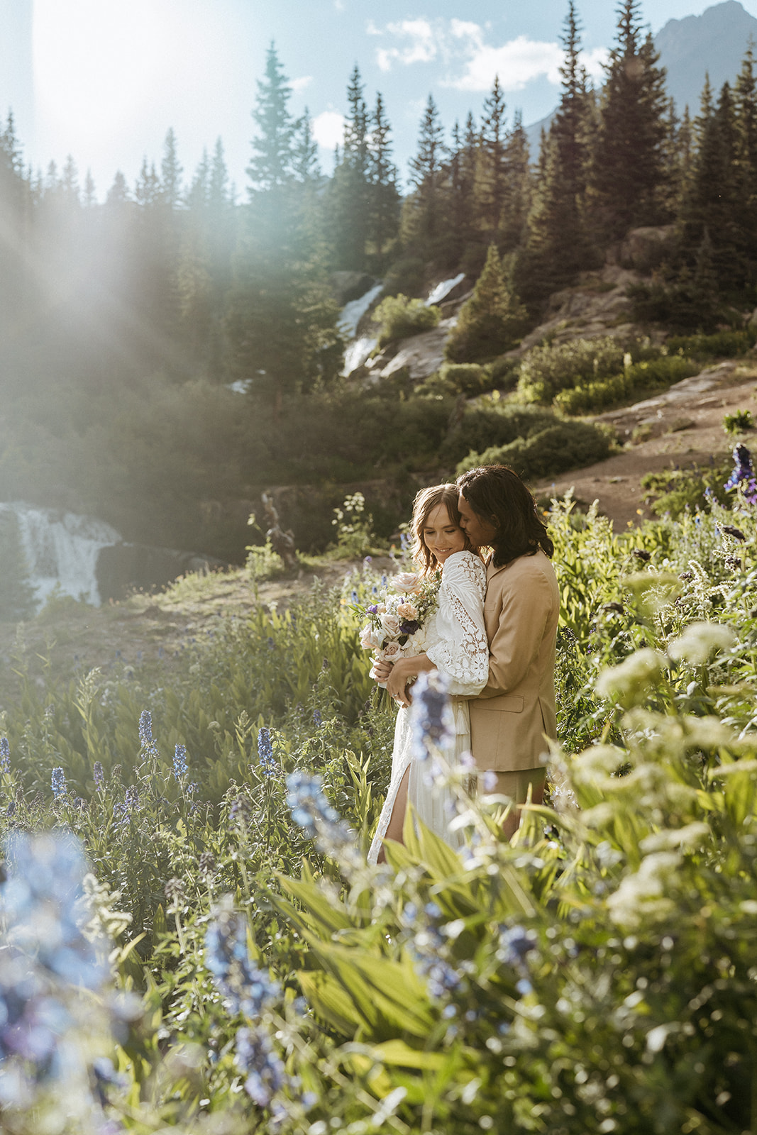 A bride leans into her groom while hiking on a mountain trail in a field of wild flowers and waterfalls at an Ouray Elopement
