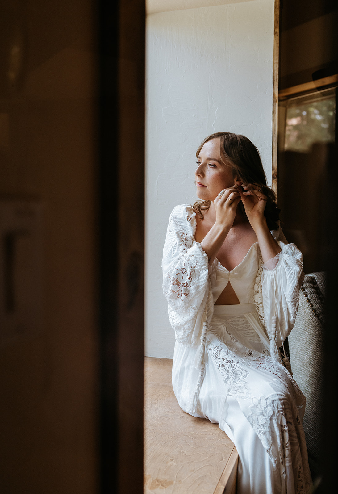 A bride sits in a window sill putting on her ear rings in a white lace dress