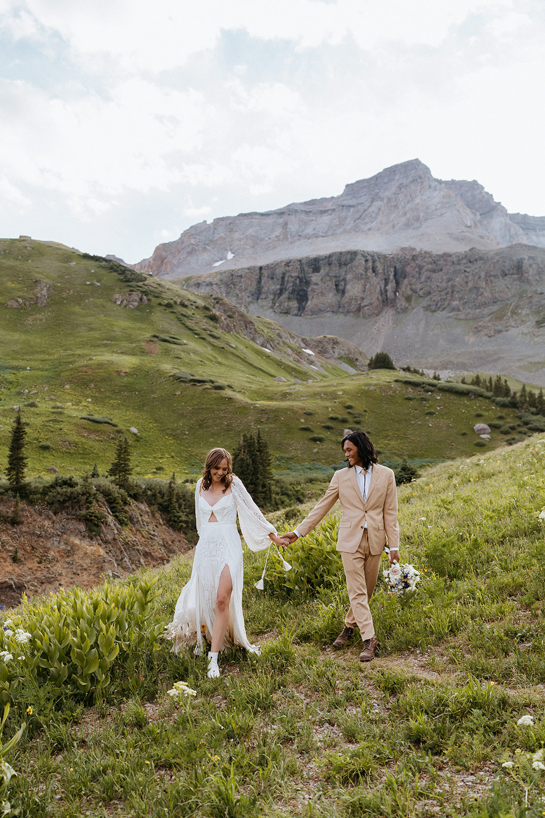 Newlyweds walk hand in hand on a mountain trail as the groom holds the bouquet and they smile for their Ouray Elopement