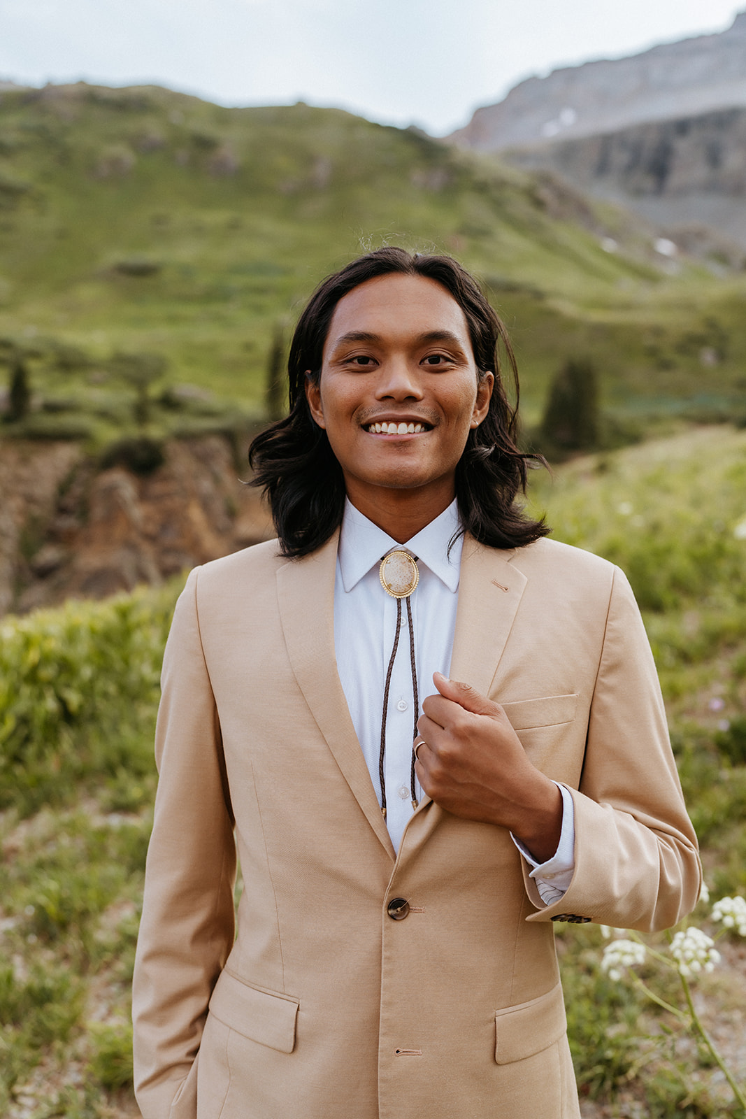 A groom in a tan suit and gold bolo tie stands on a mountain trail holding his lapel