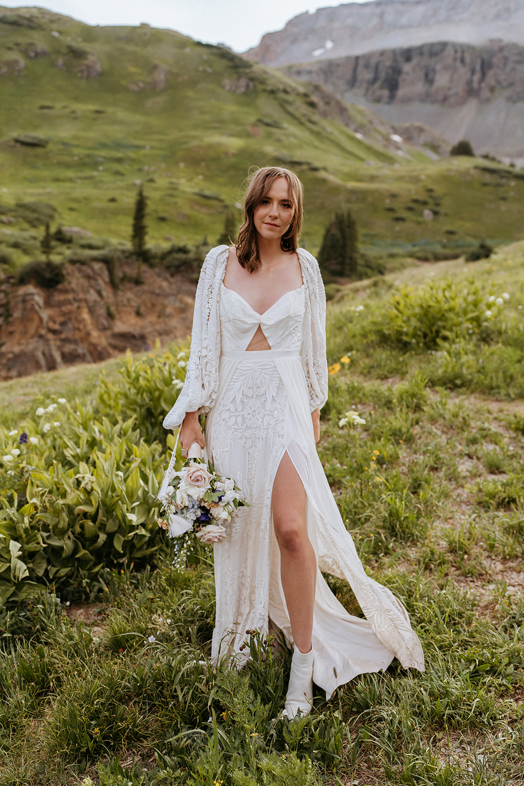 A bride in a lace dress walks through a mountain trail holding her bouquet after her Ouray Elopement