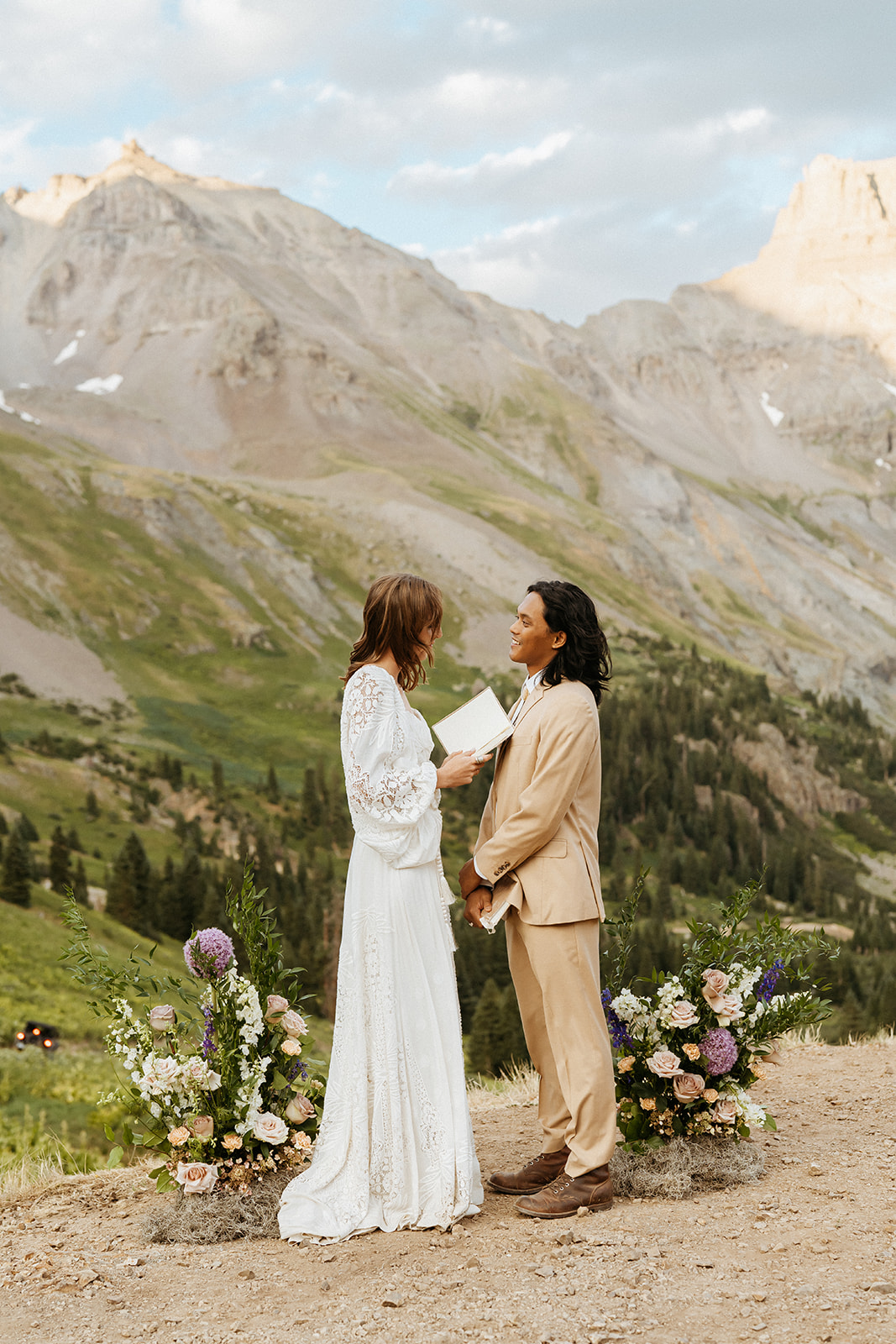 A bride in a white lace dress stands on a remote mountain trail reading her vows to her groom in a tan suit for their Ouray Elopement