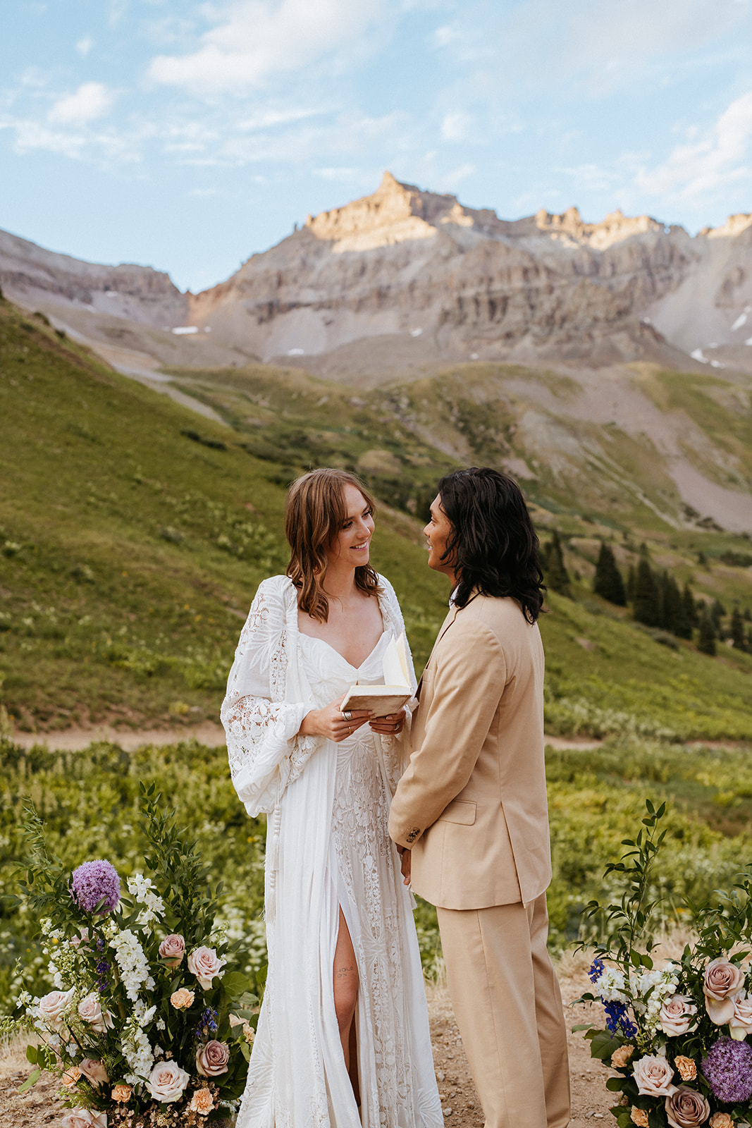 A bride in a white lace dress reads from a book on a mountain tail with two large florals to her groom in a tan suit during their Ouray Elopement