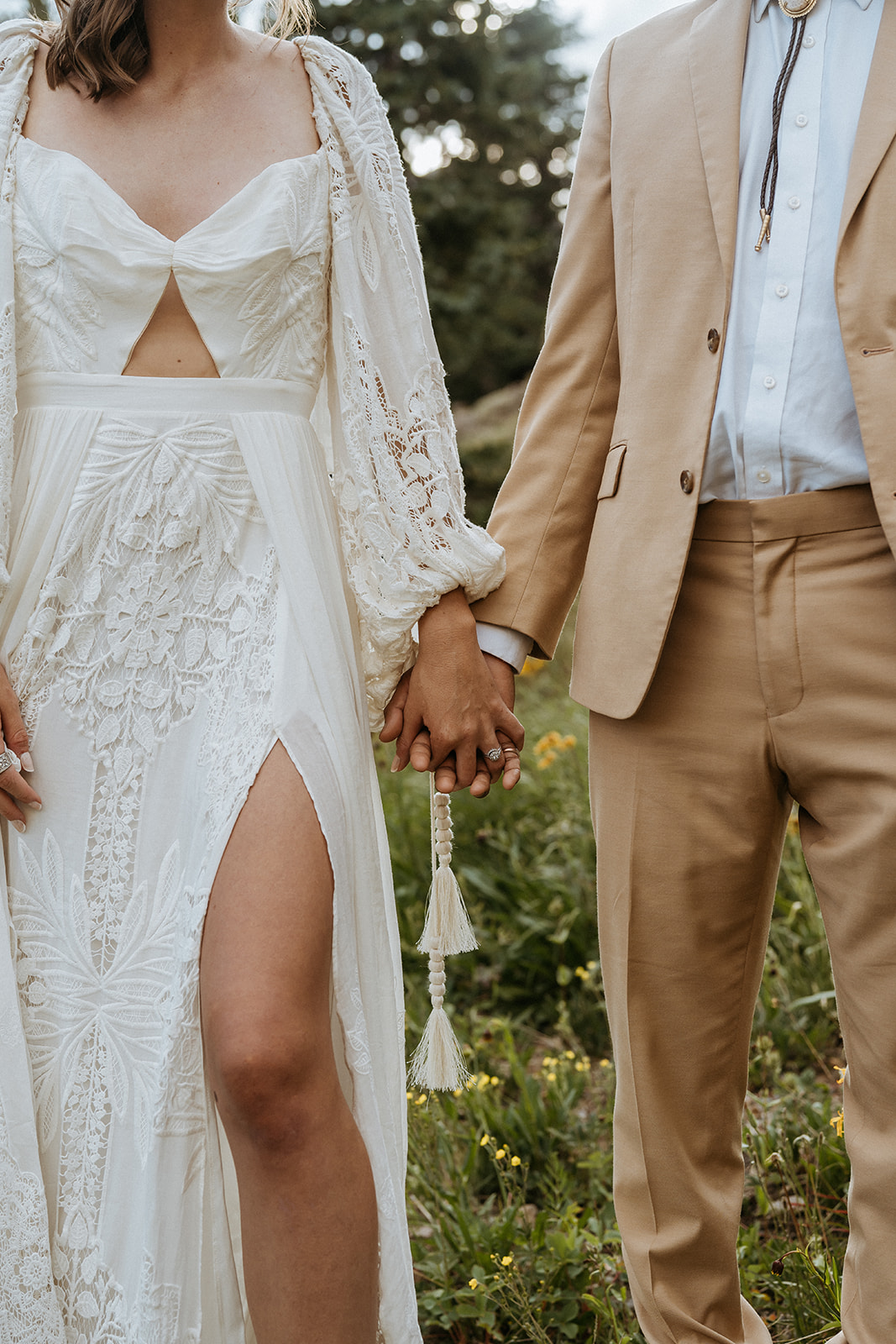 Details of newlyweds holding hands in a lace dress and tan suit on a hillside with wildflowers