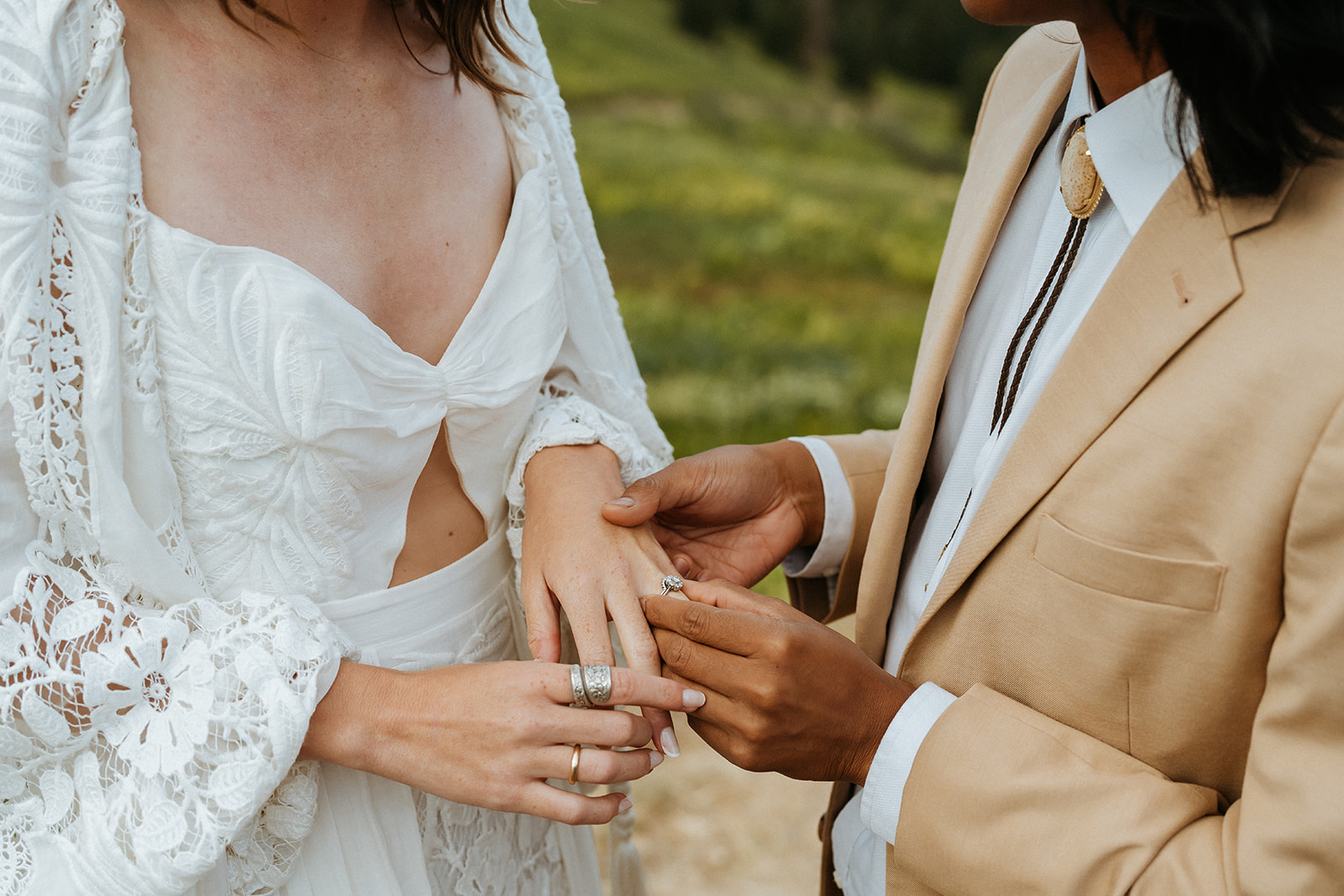 A groom in a tan suit and gold bolo tie puts the ring on his bride's finger