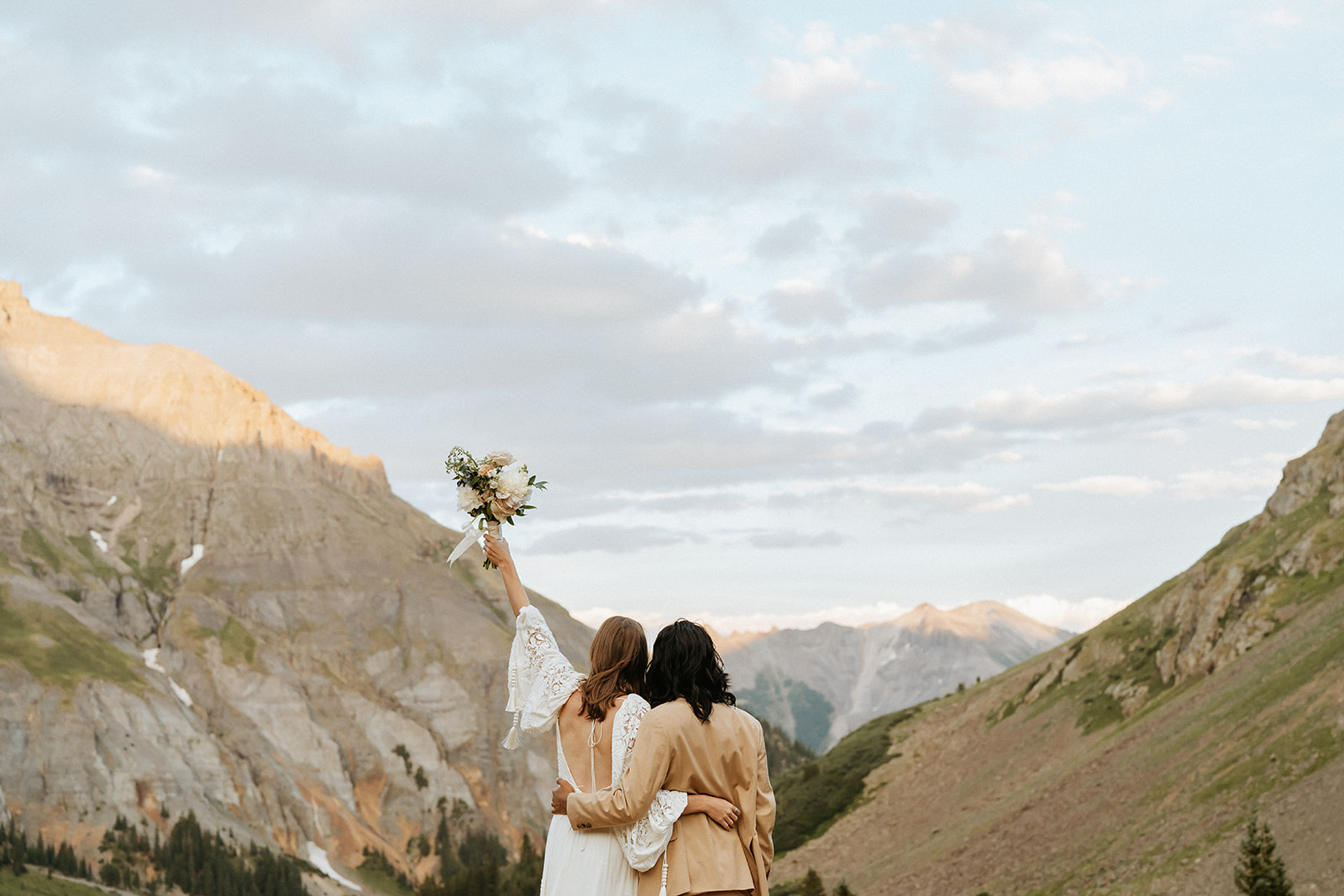 Newlyweds stand on the edge of a mountain while the bride holds up her bouquet overlooking a large valley