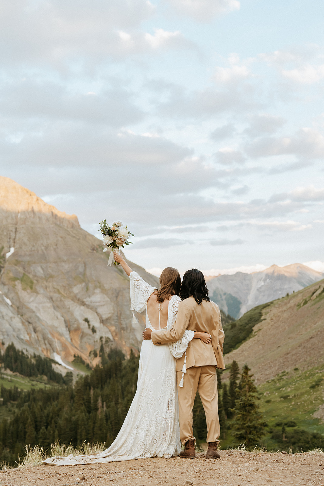 Newlyweds stand on the edge of a mountain while the bride holds up her bouquet overlooking a large valley during their Ouray Elopement
