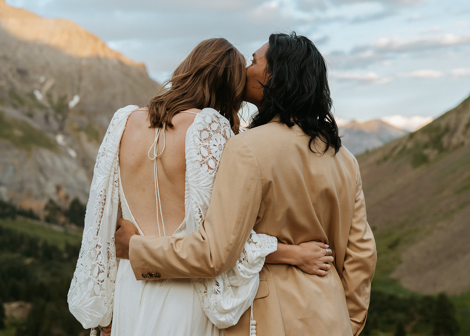 A groom in a tan suit kisses the cheek of his bride in a lace dress during their Ouray Elopement
