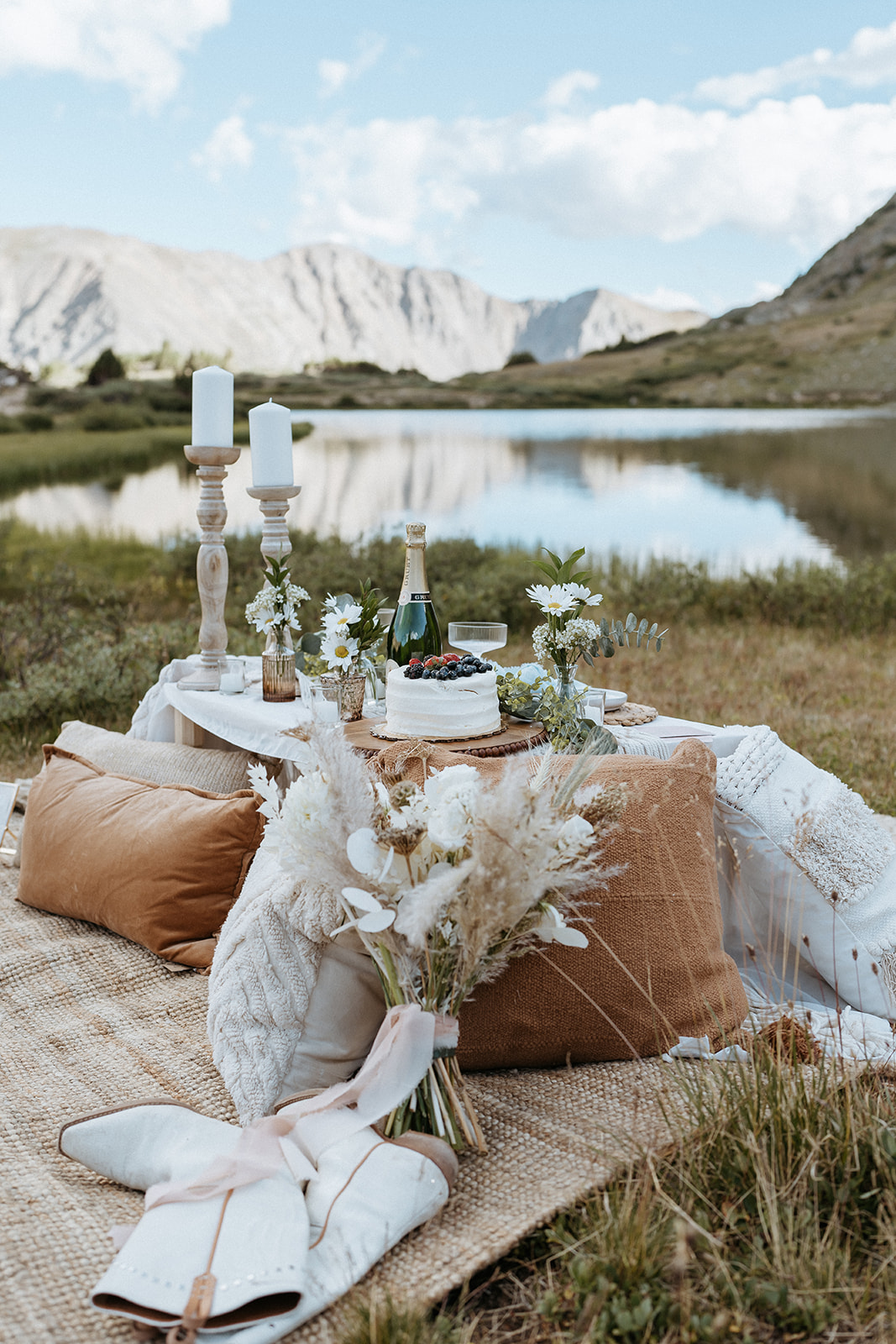 A picnic table set up by a mountain lake with pillows, cake and champagne