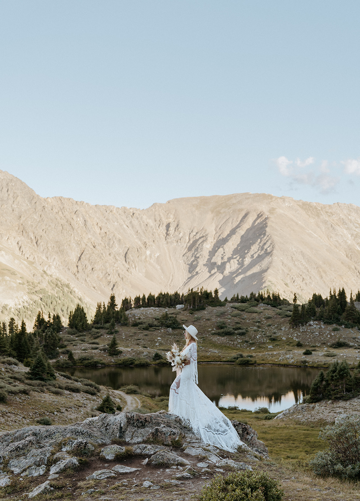 A bride in a land embroidered dress stands on a rock overlooking a lake at her Hoosier Pass Elopement