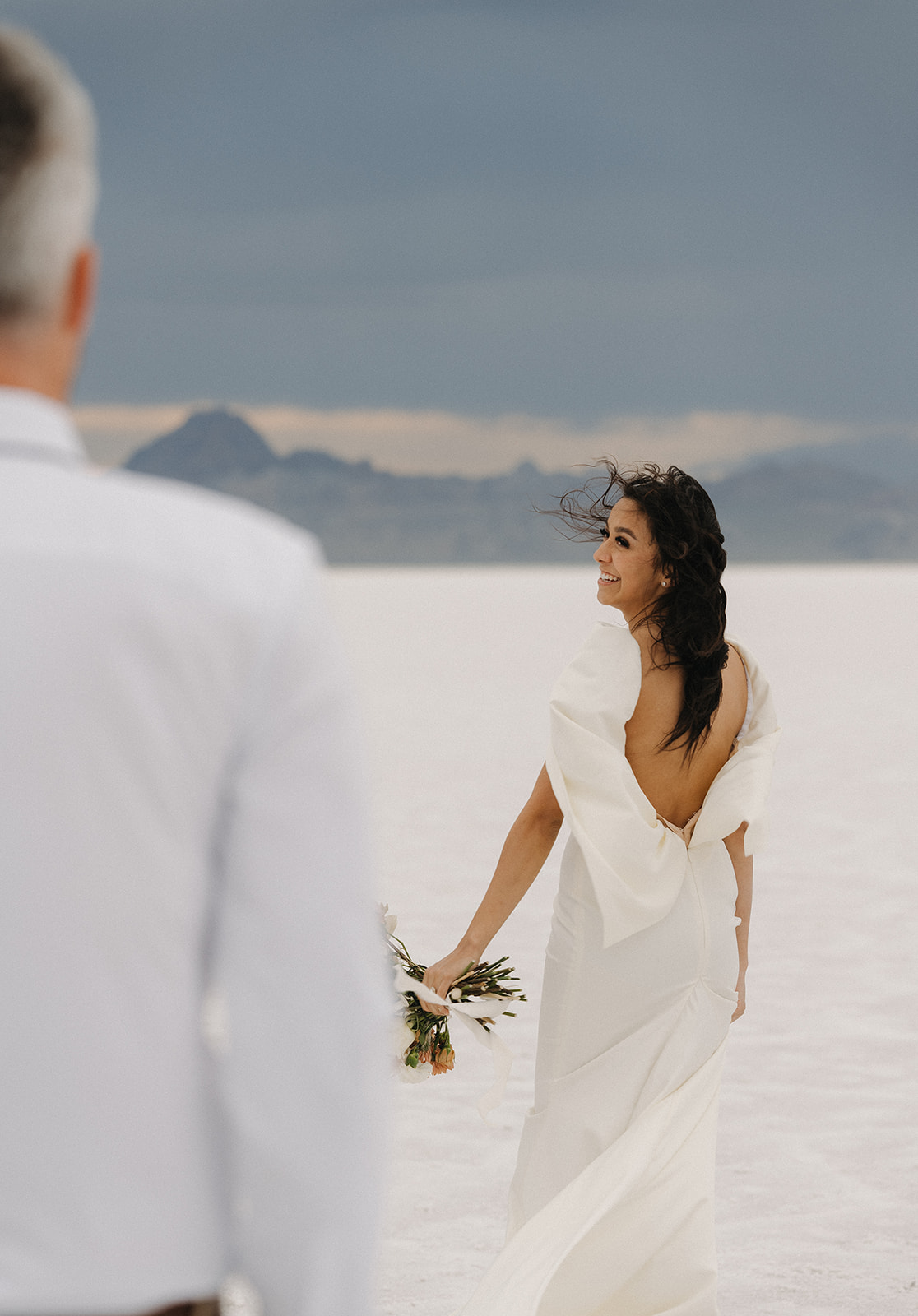 A bride looks over her shoulder to her groom while walking towards the mountains during their Salt Flats Elopement